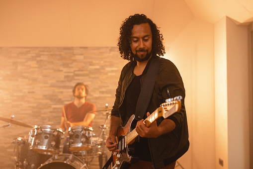A portrait of an adult black male guitarist focusing on playing the electric guitar in a beautiful studio. The musician has long curly hair and is dressed in dark clothing. He looks happy and focused. At the back of the studio there is a caucasan drummer in a red t-shirt playing the drums. The studio looks cosy and charming due to the soft warm lights.