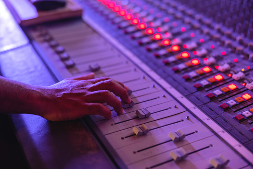 A hand of a black male audio engineer Sliding the buttons up and down on an audio mixer in a dark recording studio. There are orange lights lit on the music mixer and the recording studio is illuminated by purple neon light. The buttons on the audio mixer are white, grey, orange and red.