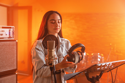 A portrait of a young adult  caucasian female vocalist preparing sing and record a song in a beautiful orange recording studio. She is putting on her headphones and standing in front to the microphone. Next to her there is recording equipment and a music stand. in the background there are drums partly visible. The musician has long brown hair and wears a denim jacket.