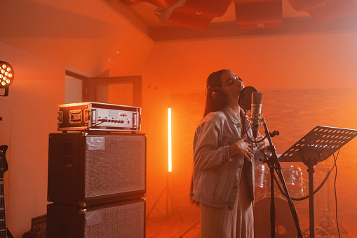 A young adult  caucasian female singer passionately recording a song in a cool recording studio. The female singer is attractive and wears casual clothes. The studio is full of audio and recording equipment. There is a microphone stand, a music stand and some drums. There is a beautiful orange light illuminating the room.