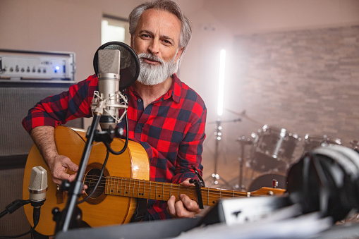 Senior Adult Caucasian Male Music Artist Looking Into A Camera While Playing An Acoustic Guitar In A Recording Studio