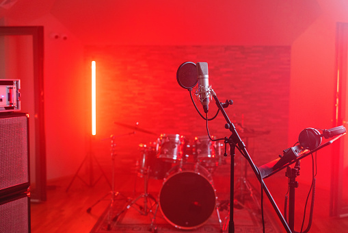 A cool small studio illuminated with a red light. The recording studio is full of sound recording and music equipment. There are microphones, music stand, headphones, loudspeakers, amplifiers and drums in this cool red music studio.