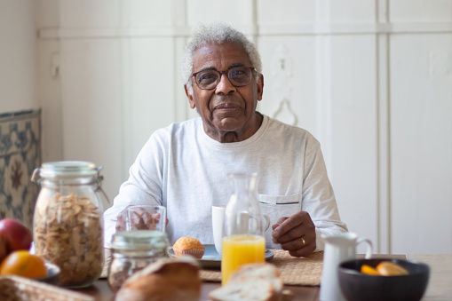 Portrait of aged man resting before breakfast in kitchen. Happy grey-haired man calmly sitting at table waiting for tea prepared and looking at camera. Support and taking care of aged people concept