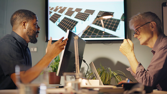 Stock image of two men, one black and the other white & mature meeting to discuss solar pale installation design… this is shown on a screen in the office.