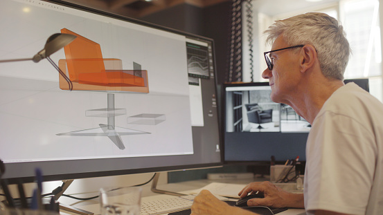 Stock image of a mature male furniture designer working with CAD to design a contemporary chair, he’s working on a large computer screen.