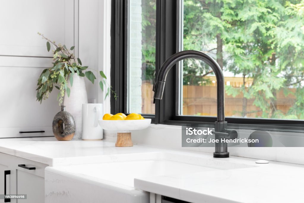 A black faucet detail shot with a white farmhouse sink. Kitchen sink detail shot in a modern, renovated kitchen with black window frames, a dark faucet, white cabinets, farmhouse sink, and cozy decor. Sink Stock Photo