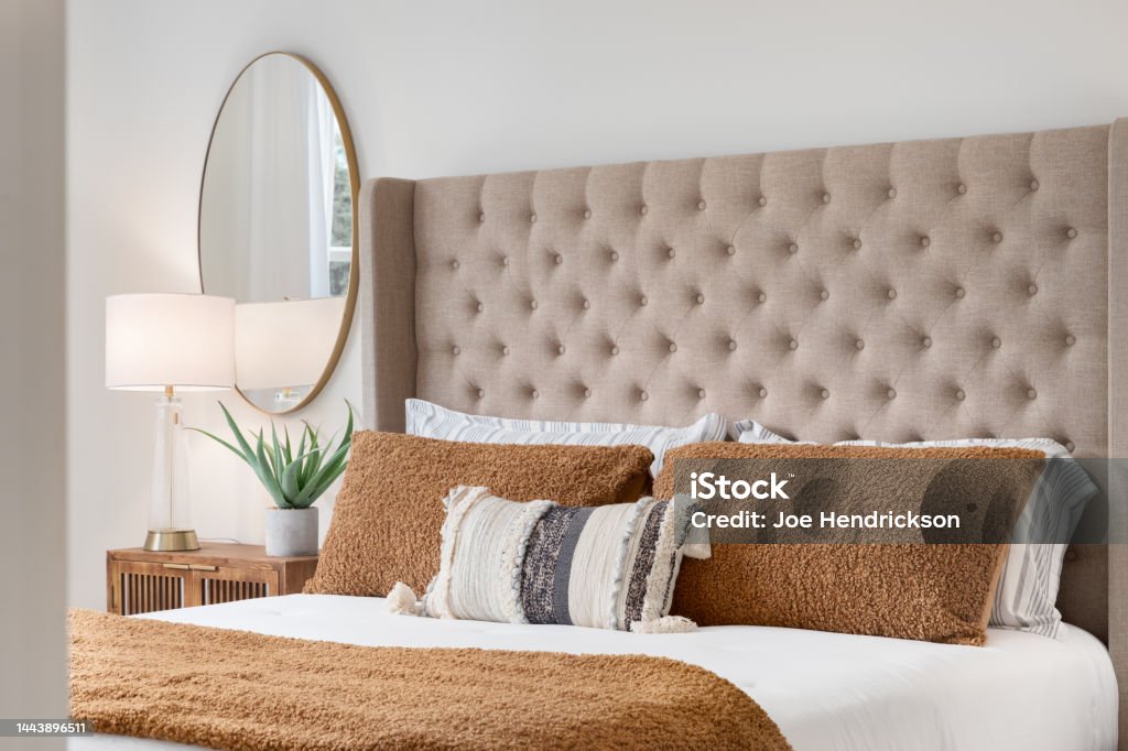 A bedroom detail with a brown headboard and bedding. A bedroom detail shot with a cushioned headboard, a gold mirror on the wall, and a plant and lamp on the nightstand. Home Interior Stock Photo