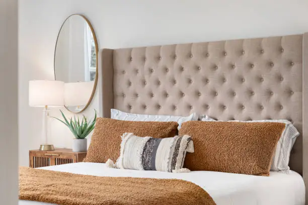 A bedroom detail shot with a cushioned headboard, a gold mirror on the wall, and a plant and lamp on the nightstand.