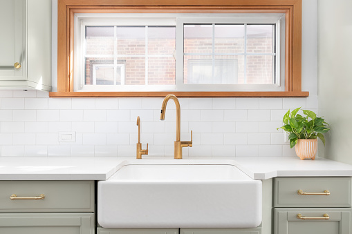A kitchen sink detail shot with a gold faucet, apron sink, subway tile backsplash, and a light green cabinet.