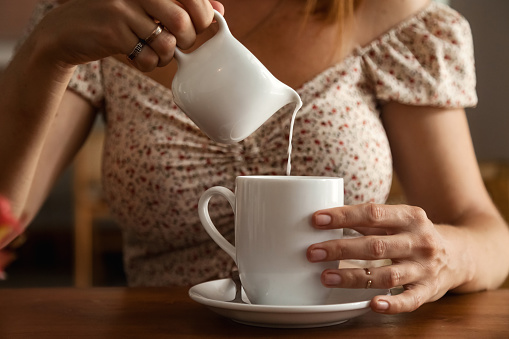 Close up middle aged woman pouring milk into cup with coffee or tea at table, resting in summer cafe with tropical flowers, sitting indoors. Travel tourism and cooking concept. Copy text space