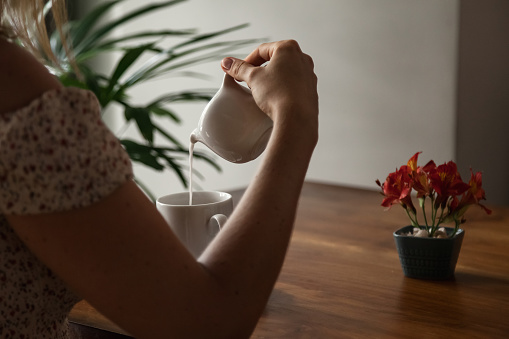 Close up rear view adult woman pouring milk into cup with coffee or tea at table, resting in summer cafe with tropical flowers, sitting indoors. Travel tourism and cooking concept. Copy text space