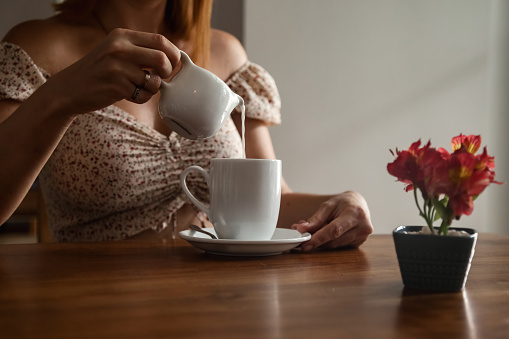 Close up middle aged woman pours milk into mug with coffee or tea at table, relaxing in summer cafe with tropical flowers, resting indoors. Travel tourism and cooking concept. Copy text space