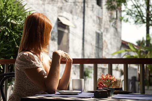 Middle aged woman sitting at table with coffee or tea cup and flowers, in summer outdoor terrace cafe with tropical green plants, resting and looking away. Travel tourism vacation concept. Copy space
