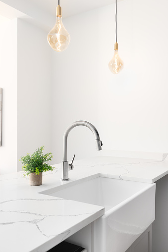 A kitchen sink detail shot with glass pendant lights hanging above the white granite countertop, apron sink, and chrome faucet.