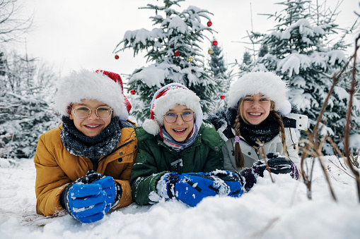 Three kids have decorated a Christmas tree outdoors on a winter day. They are lying on the front on snow and smiling at the camera.\nCanon R5