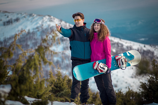 Happy young couple looking at the horizon, the boy is pointing with his finger, the girl is holding a snowboard in her hand.