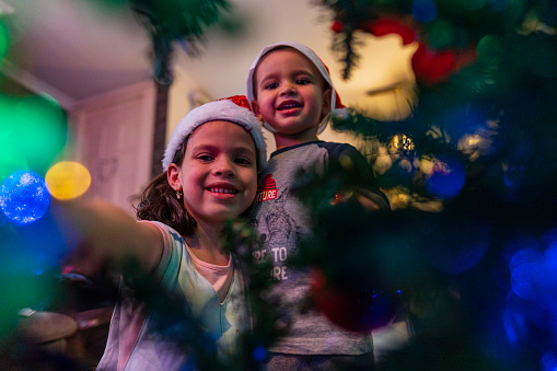 Latino brothers and sisters, an average age 7-year-old girl with short brown hair dressed very well and a 3-year-old Latino boy, both with Christmas hats, are inside their house that is fully decorated with Christmas lights and decorations, they are at the foot of their tree together placing the decorations and enjoying a beautiful moment of brothers