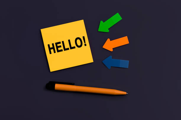 istock Hello salutation or greeting word to welcome someone or initiate a conversation. Communication concept, introduction. Yellow square sticky note, pen and arrows of paper on dark color background 1443911847