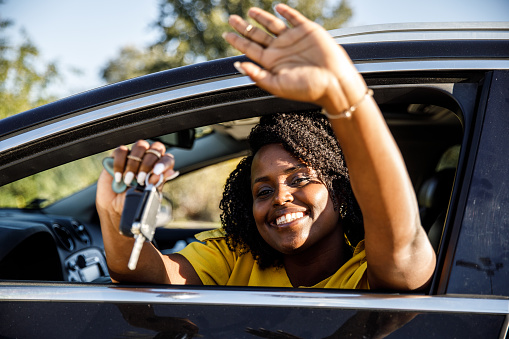 Portrait of happy young woman sitting in the driver's seat of her new car, waving through the open car window and showing you off her new car keys while joyfully smiling.