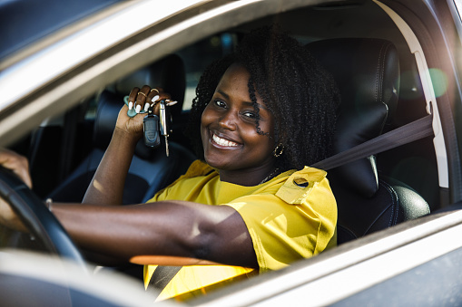 Portrait of happy young woman sitting in her new car and showing off new keys that she is holding while smiling at camera through the car window.