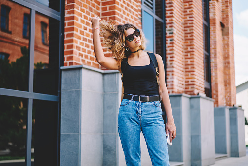 Confident female in casual clothes and sunglasses tousling hair while standing near brick wall on street and looking away on sunny day