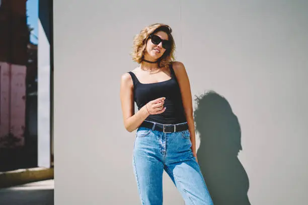 Young smiling content female with brown hair in sunglasses wearing denim and choker posing while standing on gray background on sunny street
