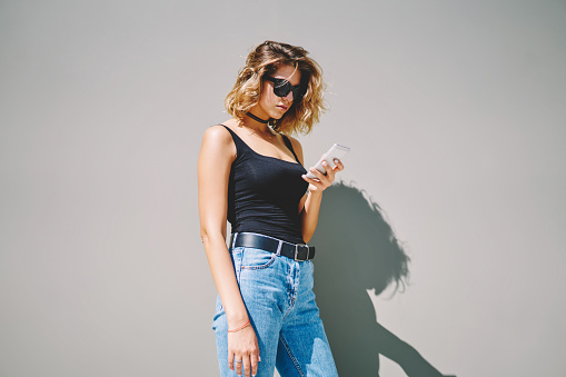COncentrated female in jeans and sunglasses text messaging on smartphone and looking at screen while standing on gray background on sunny day