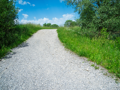 View along an uphill gravel road in a hilly landscape surrounded by grass and small trees in front of a blue summer sky with small white clouds.