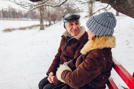 Senior family couple sitting on bench outdoors during snowy winter weather. Elderly retired people chatting in park. Valentine's day