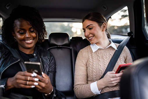 Front view of two diverse young women sitting in the back seat of a crowdsourced taxi and using their smart phones to pass the time while riding to their location.