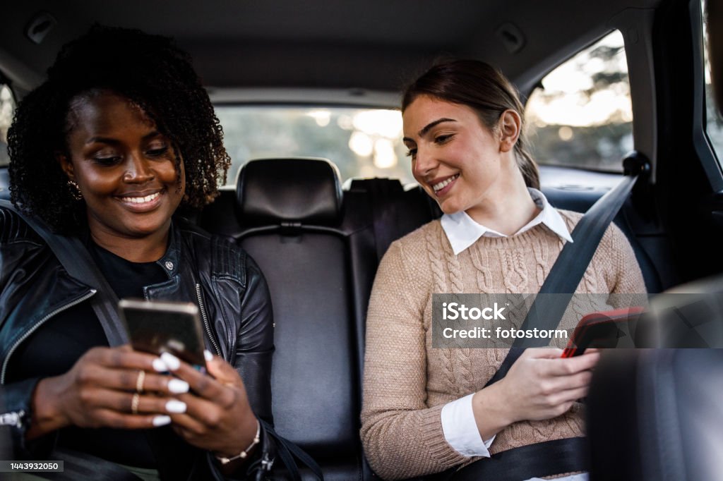 Two young women riding in the back seat of a taxi, using their smart phones to pass the time Front view of two diverse young women sitting in the back seat of a crowdsourced taxi and using their smart phones to pass the time while riding to their location. Car Pooling Stock Photo