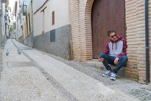 Young man sitting on the step of the house outdoors, serious and reflective looking into the distance, on a street in the historic district of Granada. Outdoor waiting concept.