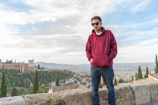 Young man posing at the viewpoint of San Nicolas in Granada, with red sweatshirt in the foreground, in the background the Alhambra and the city of Granada.