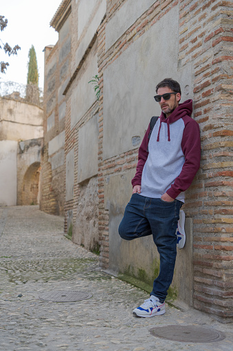 Young man model with a sweatshirt and standing leaning against the wall, poses in a street of the Albaicin in Granada