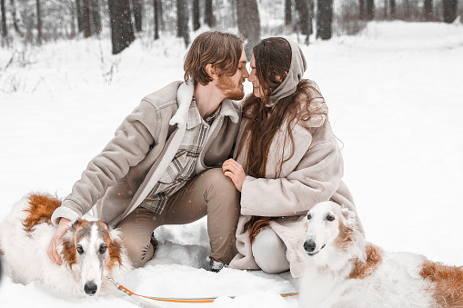 Love romantic young couple girl, guy in snowy cold winter forest walking with pet, dog of hunting breed russian borzoi. Sighthound, wolfhound owner. Having fun, laughing. Stylish fur coat, woolen hat.