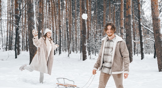 Love romantic young couple girl,guy in snowy winter forest sledding,playing snowballs.Walking with sleigh in stylish clothes, fur coat,jacket, woolen shawl,bonnet.Snow lovestory.Romantic date,weekend.