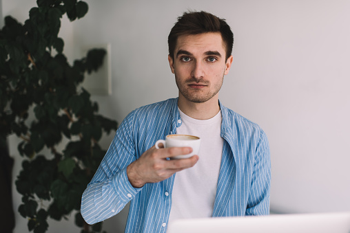 Half length portrait of young Caucasian man 20 years old with hot caffeine beverage posing indoors, trendy dressed male student in smart casual clothing holding coffee cup and looking at camera