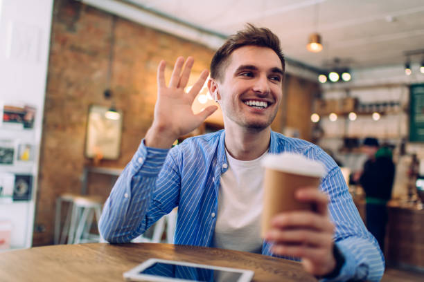 istock Cheerful hipster guy with takeaway cup in hand waving while waiting best friend in cafeteria interior, happy male millennial in electronic earbuds listening positive audio records from touch pad app 1443943253