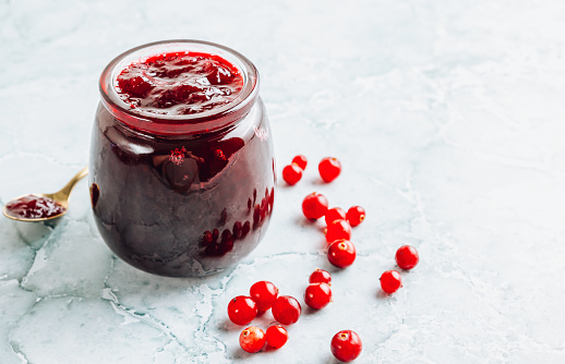Jar of tasty cranberry jam on light marble background. Preserving cranberry sauce. Selective focus