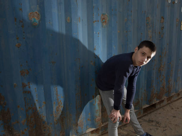attractive and stylish fair-haired young caucasian guy posing with a serious face dressed in a sweater and jeans in an industrial area against a blue iron wall and a blue cloudy sky. lower angle