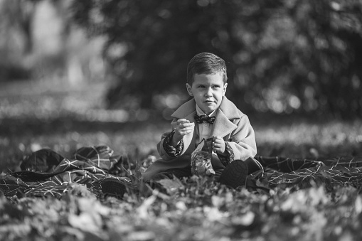 Portrait of a handsome little boy sitting in park and eating snacks on a beautiful autumn day. Black and white.