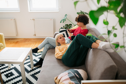 Two young adult women relaxing at home, lying on a sofa together and cuddling