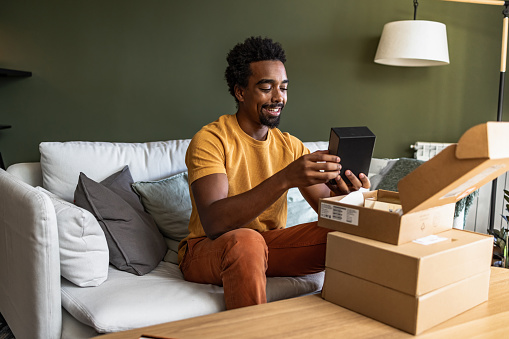Young  African American man feeling happy while opening his online purchase at home.  He is sitting on the couch in the living room. Cardboard boxes are on the table.