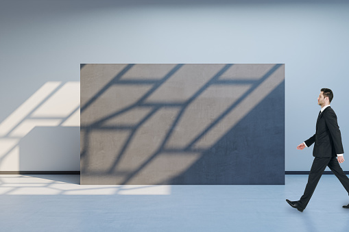 Side view of blurry man walking in spacious gallery interior with empty poster and sunlight shadow on concrete wall. Mock up