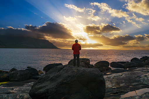 Hiker standing on Tungeneset beach at sunset with mountains under heavy clouds in the background, Senja Island, northern Norway