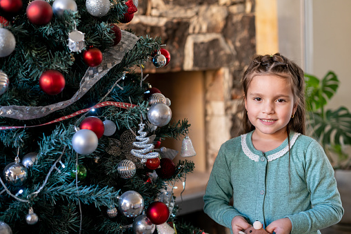 Cute Latin American little girl standing next to the Christmas tree while facing camera smiling - Celebration concepts