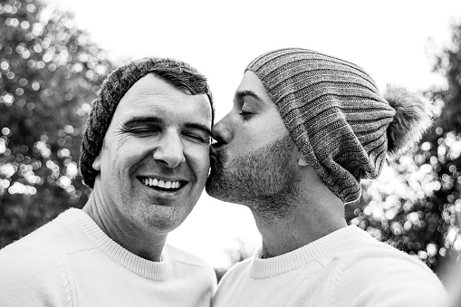 Happy mature gay men couple taking selfie kissing in winter time outdoor - Lgbtq love concept - Black and white editing