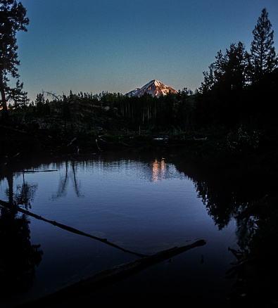 Engineer Mountain is seen in the distance. the trees are casting dark shadows in the lake and are in silhouette. Reflection of the trees and mountain are seen in the lake.