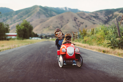 A boy wearing racing goggles races his toy race car with his support dog on a small road. This is a road trip for the soul. Image taken in Utah, USA.