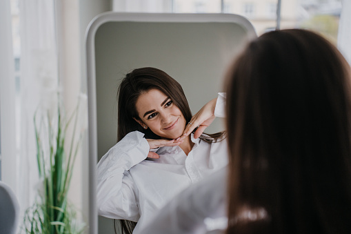 Hispanic gorgeous young woman in white shirt at home looks at mirror, smiling, satisfied after cosmetology procedure, skin care, health and beauty concept. Beauty and healthy lifestyle.